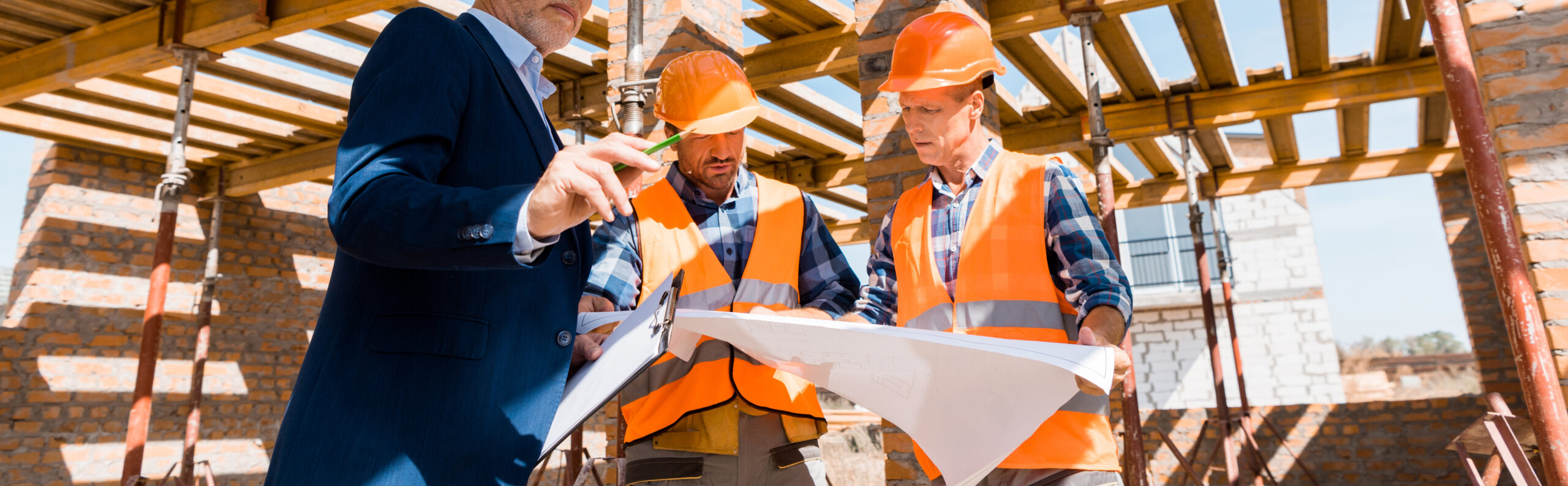 An owner and two contractors in hard hats reviewing plans beneath wood roof trusses